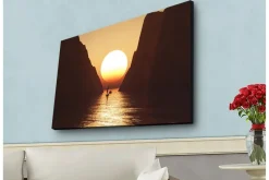 Canvas-taulu LED-valaistuksella - Upea auringonlasku kapean vuonon yllä purjeveneen siluettina Oranssi / Ruskea / Kulta